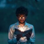 A man with glasses reads an illuminated book outdoors during twilight.